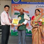 A man in a pale pink shirt hands a certificate to a man in a green kurta on stage while a woman in a beige sari with a bouquet stands nearby and a student watches on the left